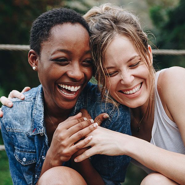 Two women sitting close together outdoors, smiling and laughing joyfully. One wears a denim jacket, the other a white tank top. Their arms are around each other, expressing happiness and friendship.